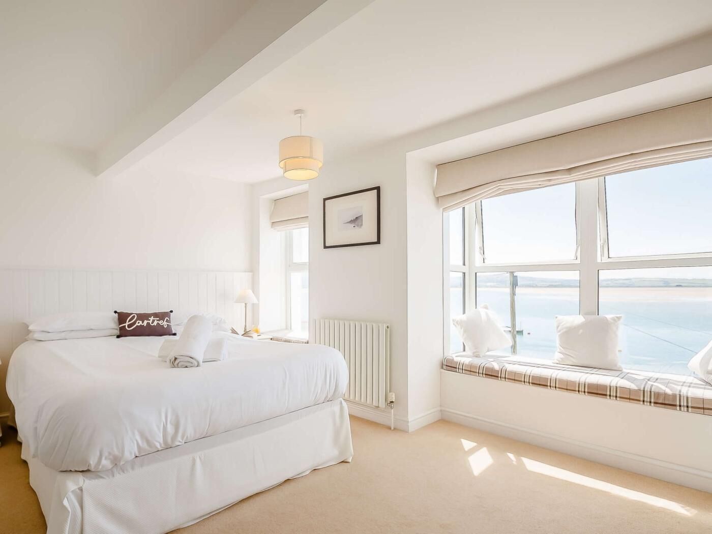 A hotel room with a large bed, a window seat, a radiator, and a hanging light fixture.