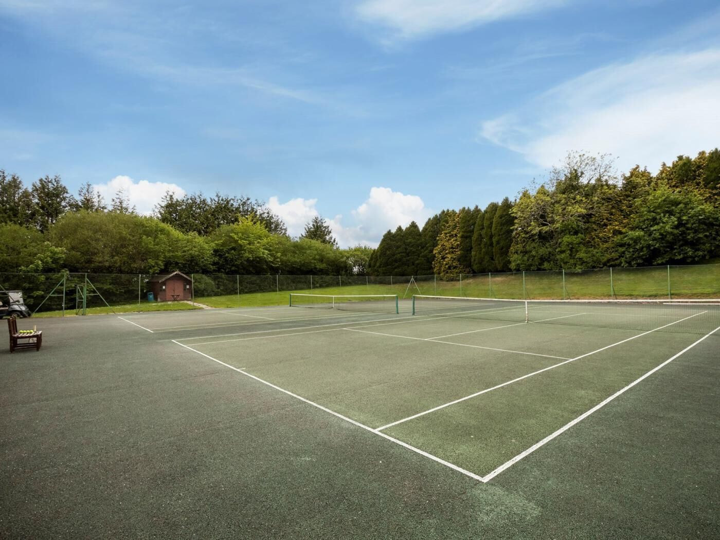 En udendørs tennisbane med net, omgivet af træer og en mindre bygning.