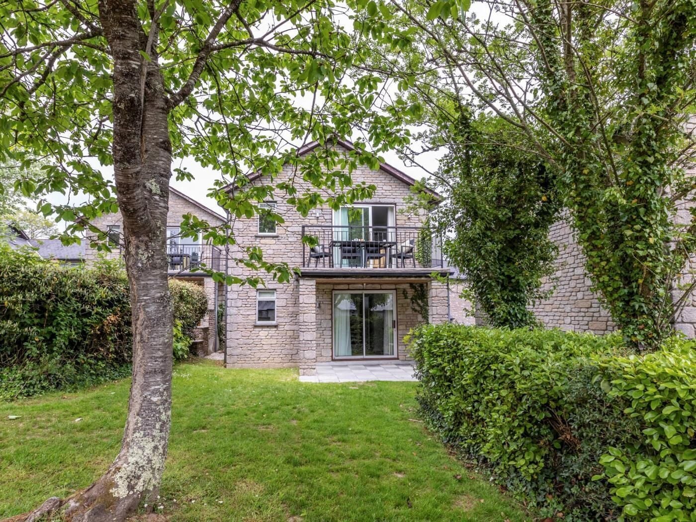 Une maison en pierre avec un balcon, une cour gazonnée et un arbre.