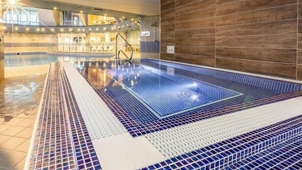 An indoor swimming pool with a tiled floor, a wooden wall, and a sunlit area with lounge chairs.
