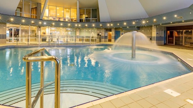 Piscina interior com um elemento central de água, rodeada por um design arquitetônico moderno com fachada de vidro e um mezanino.