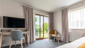 A hotel room with a bed, a desk with a TV, a chair, a small table with a cup, and a view of greenery outside. - 2 Bed in Saltash (oc-87704) (Saint Mellion)