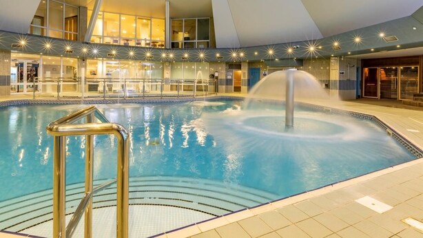 Piscina interior com uma fonte central, cercada por um design arquitetônico moderno, teto alto e paredes de vidro.