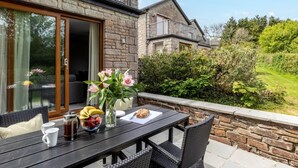 A patio with a table set for breakfast, a stone wall, and a view of a house with a balcony. - 2 Bed in Saltash (oc-87706) (Saint Mellion)