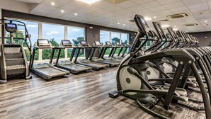 Un gimnasio con cintas de correr, elípticas y una máquina de cardio, con ventanas amplias y piso de madera.