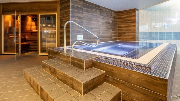Un área de piscina interior moderna con cerramiento de cristal, escalones de madera y suelo de baldosas.