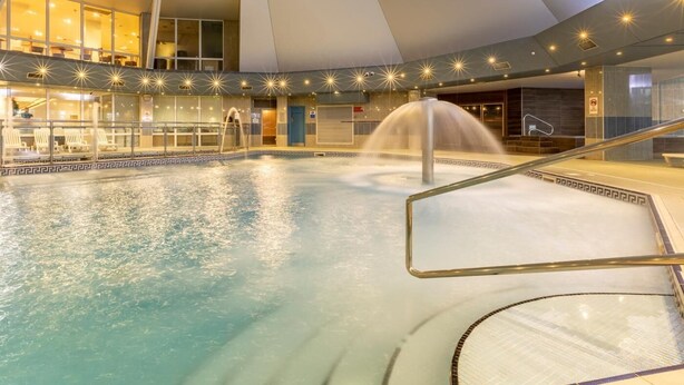 En inomhuspool med en central fontän, omgiven av ett glasräcke och med ett geometriskt mönster i taket.