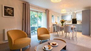 A modern living room with two armchairs, a coffee table with food, a dining area with chairs, and a kitchen in the background. - 2 Bed in Saltash (oc-87713) (Saint Mellion)
