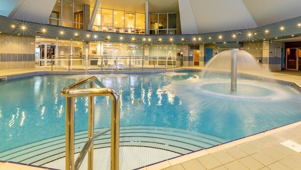 An indoor swimming pool with a fountain, surrounded by a modern architectural design with a glass enclosure and a seating area. - 3 Bed in Saltash (oc-87711) (Saint Mellion)