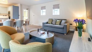A modern living room with a grey sofa, two armchairs, a coffee table, and a dining area with a table and chairs. - 2 Bed in Saltash (oc-87705) (Saint Mellion)