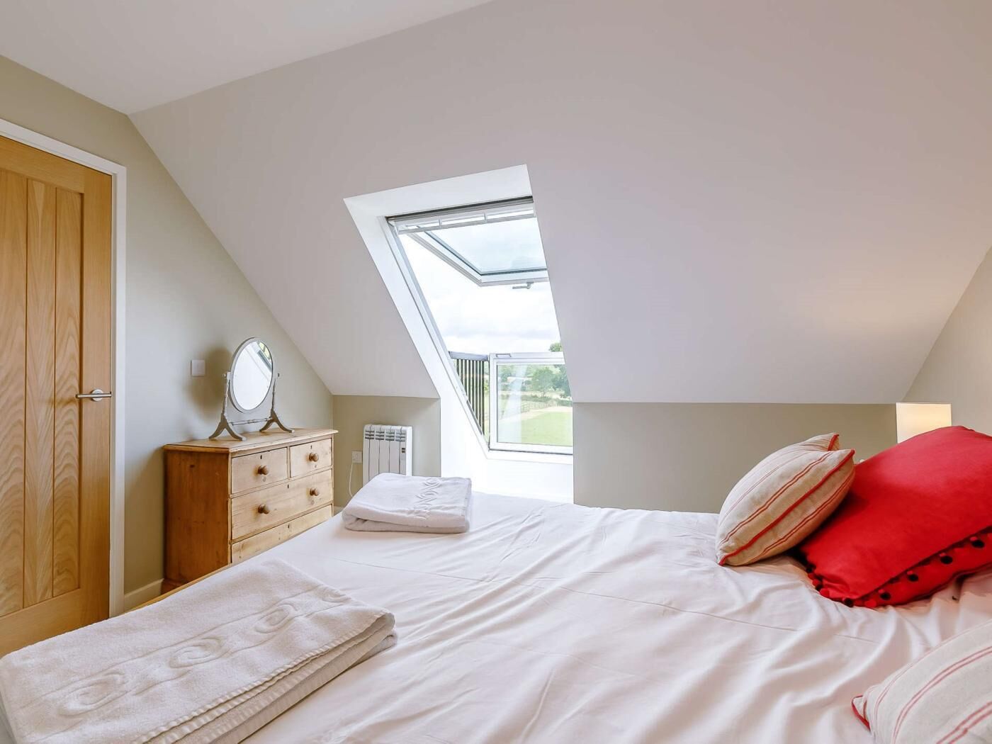 Une chambre avec un puits de lumière, une armoire en bois, une table de chevet avec un miroir, un lit avec une literie blanche et un oreiller rouge.