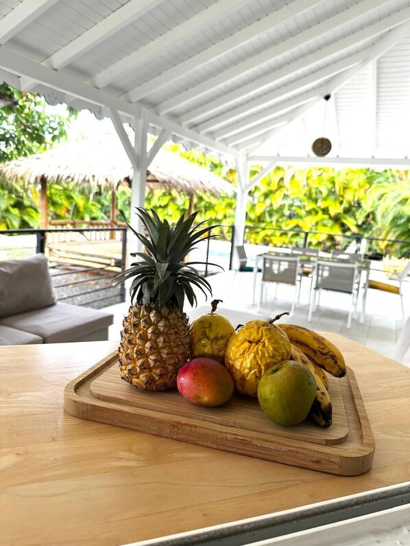Une table en bois sur laquelle est disposée une assiette de fruits, le tout placé sous une pergola blanche, et entouré d’une végétation luxuriante.