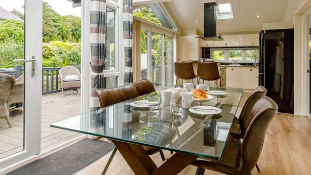 A modern dining area with a glass table, leather chairs, and a view of a garden through large windows. - 2 Bed in Barnstaple (oc-87360) (Kentisbury)