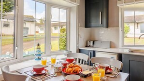 A breakfast table set with red bowls, plates, and glasses of orange juice, with a view of a residential area through large windows. - 2 Bed in Brixham (oc-88279) (Brixham)