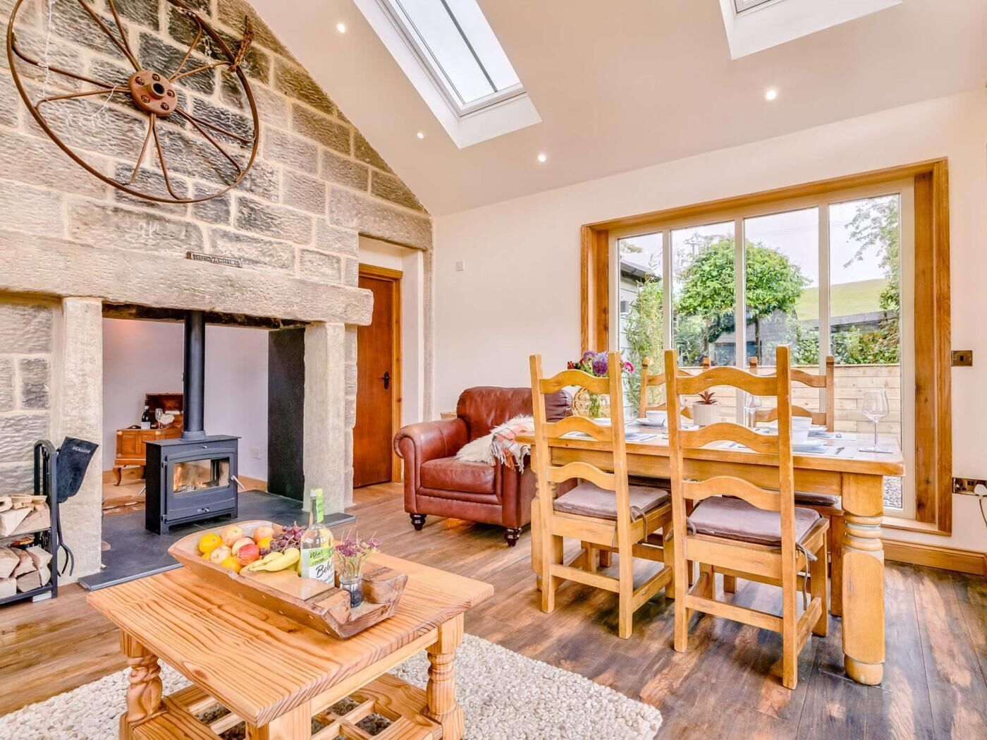 A cozy room with a stone fireplace, a dining table with chairs, a leather sofa, and a large window with a view of greenery.