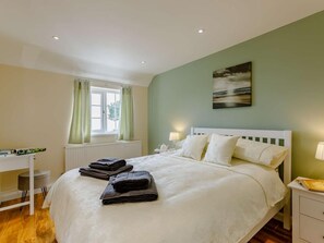 A neatly made bed with pillows and folded towels, a bedside table with a lamp, a window with curtains, and a painting on the wall. - 1 Bed in Cinderford (oc-88272) (Cinderford)