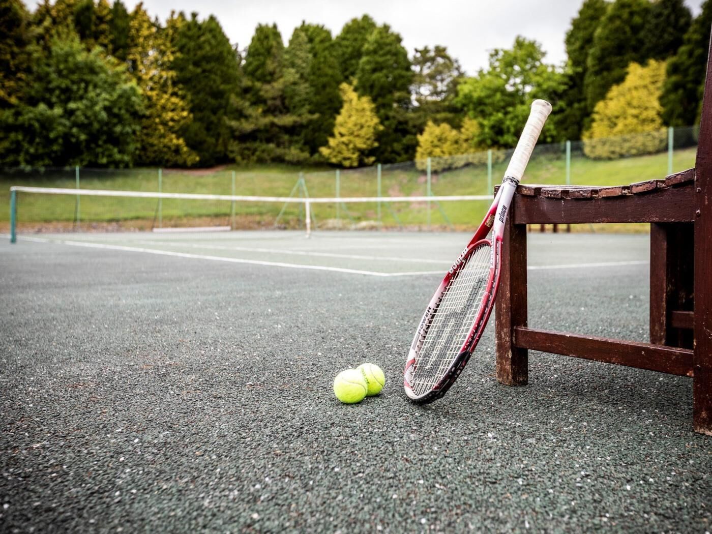 A tennis court with a net, a tennis racket leaning against a bench, and two tennis balls on the ground.
