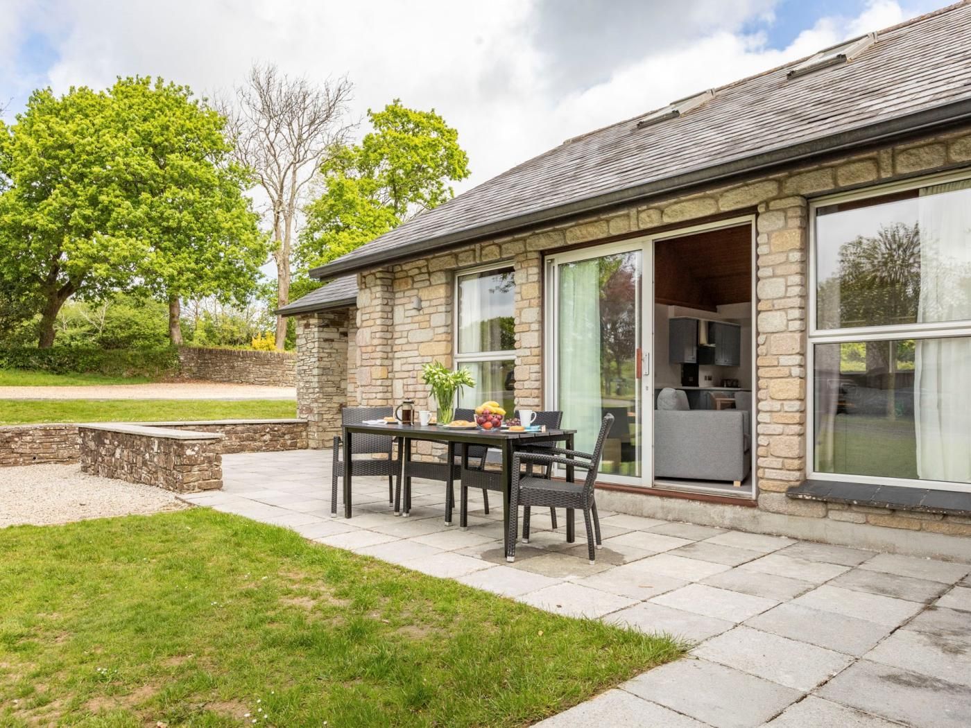 A stone building with a patio area featuring a table and chairs, a stone wall, and a grassy lawn.