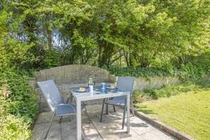 An outdoor patio with a table and chairs, surrounded by greenery and a stone wall.