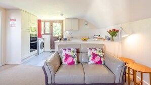 Een ruimtelijk ingerichte leefruimte met een kitchenette, een bank met bloemkussens en een klein houten tafeltje.