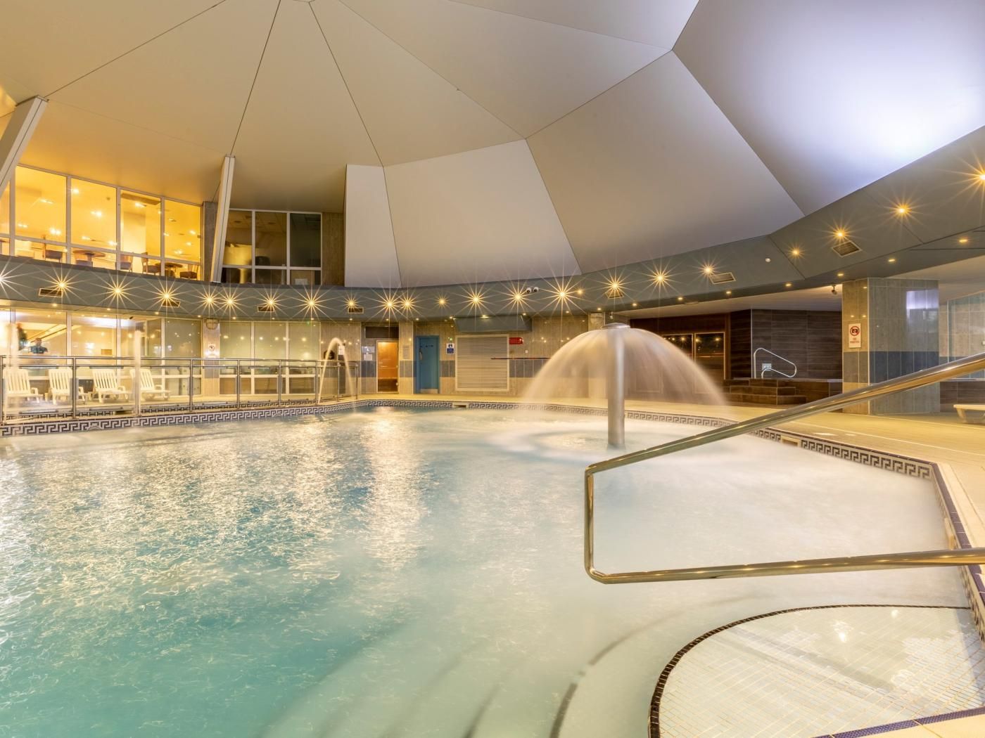 An indoor swimming pool with a central fountain, surrounded by a glass railing and a geometric ceiling design.