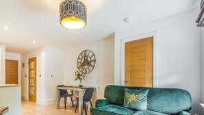 Een woonkamer met een groene bank, een eettafel met stoelen, een wandklok en een hanglamp.