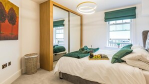 Een slaapkamer met een bed, een grote spiegel, een nachtkastje met een schaal fruit, een rieten mand en uitzicht op het buiten via een raam.