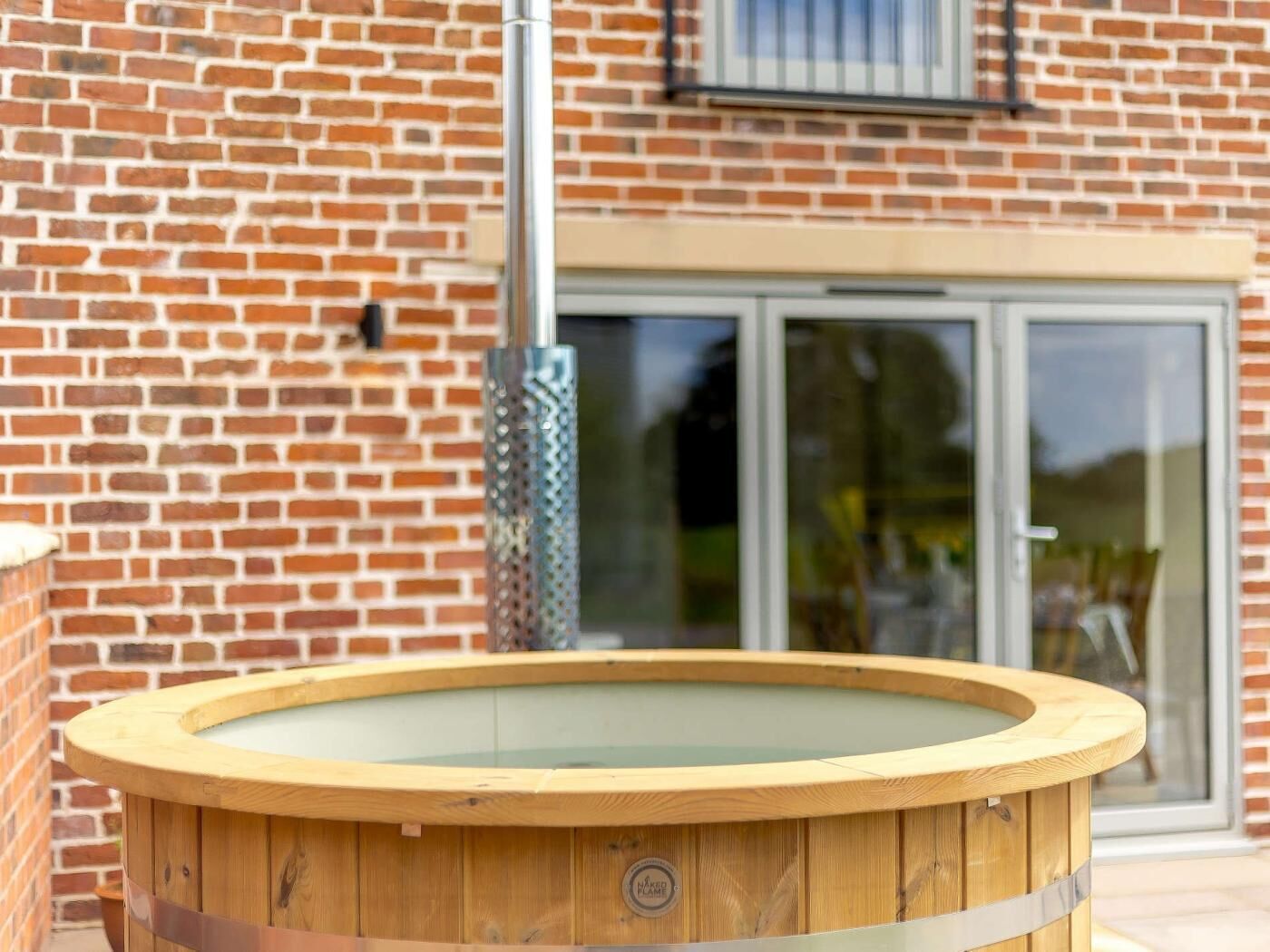 Un bain à remous en bois, surmonté d’une paroi en verre, installé devant un mur de briques, avec une porte-fenêtre coulissante en arrière-plan.