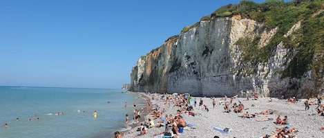 Ein belebter Strand mit Menschen, die sonnenbaden und schwimmen, ein felsiger Klipp und blauer Himmel.