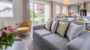 A modern living room with a grey sofa, a beige armchair, and a dining area with a kitchen in the background. - 2 Bed in Saltash (oc-87715) (Saint Mellion)