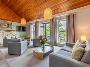 A modern living room with a stone fireplace, grey sofas, a round coffee table, and large windows with curtains. - 3 Bed in Saltash (oc-87718) (Saint Mellion)