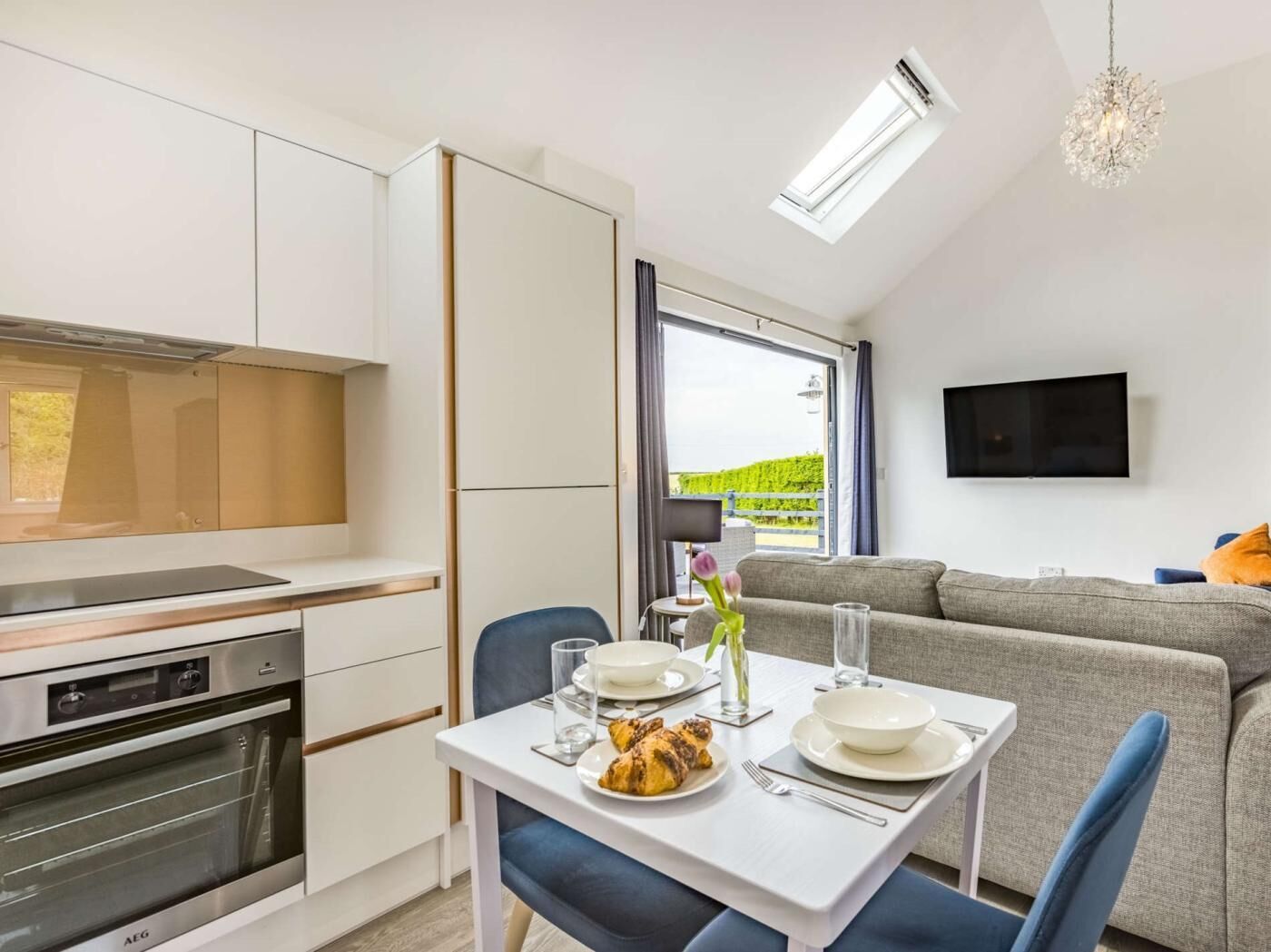 A modern kitchen with a built-in oven, a dining table set for two with plates and glasses, and a grey sofa in the living area with a TV mounted on the wall.
