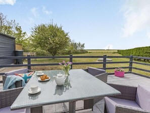 Eine Terrasse im Freien mit einem Glastisch, Korbmöbeln und Blick auf ein Feld und Bäume.