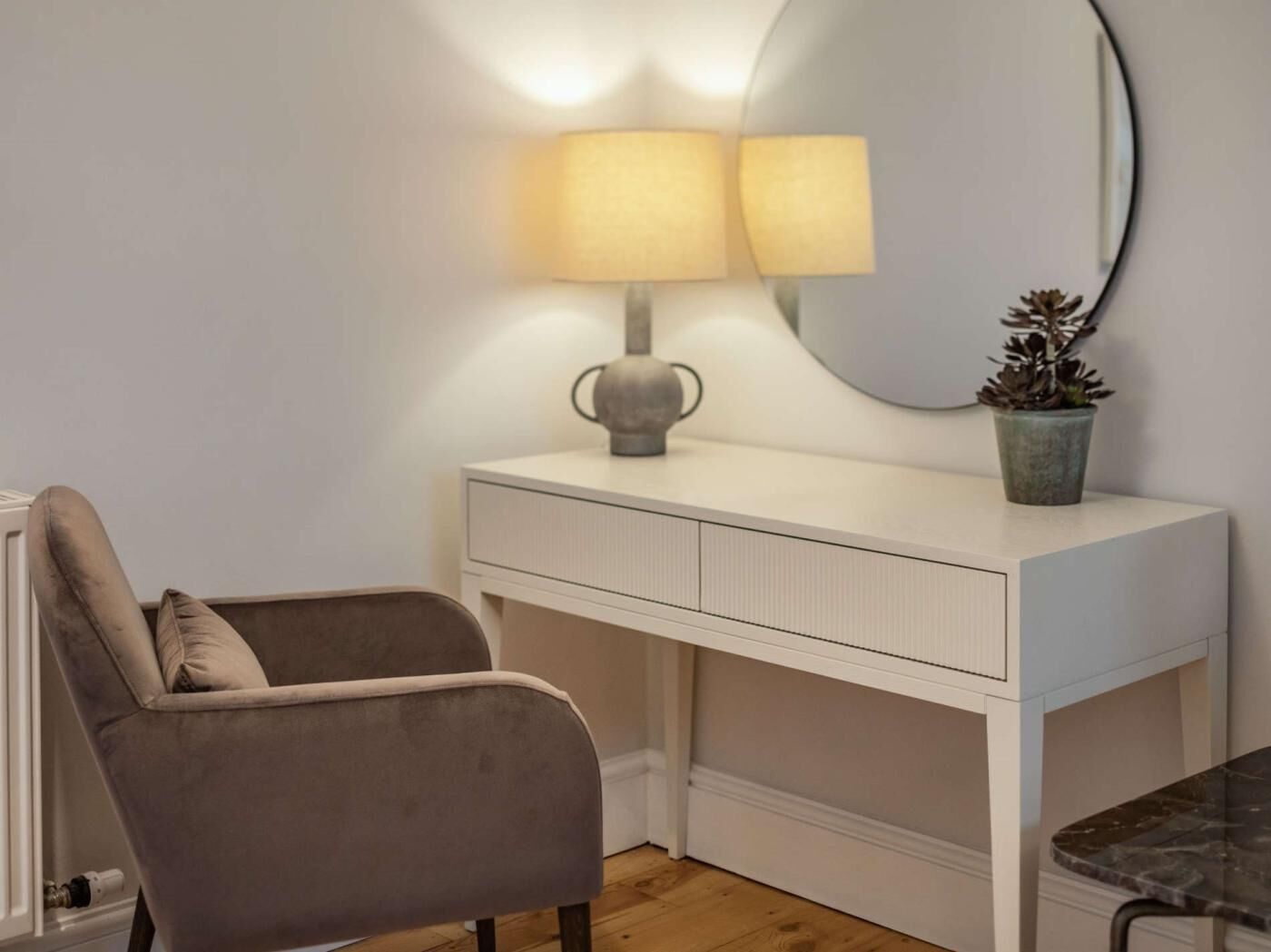 A white vanity with a round mirror, a lamp, and a potted plant.