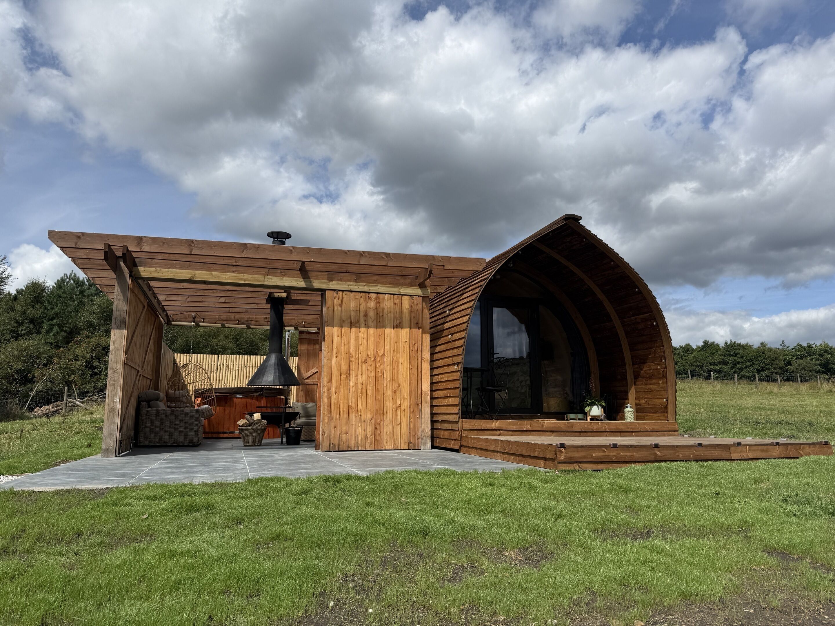 A wooden cabin with a chimney, a covered seating area, and a large window.