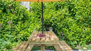 Una taula de picnic de fusta amb un parasol groc, envoltada d'arbustos verds i frondosos.