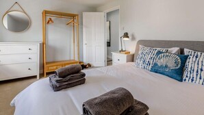 A bedroom with a bed, a bedside table with a lamp, a wardrobe, a mirror, and a hanging coat rack. - 3 Bed in Budleigh Salterton (oc-88004) (Otterton)