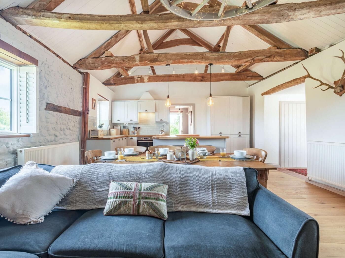 A rustic interior with exposed wooden beams, a dining area with a table set for four, and a sofa with pillows.