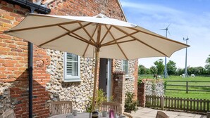 Een ronde houten tafel met stoelen en een grote parasol, voor een stenen muur met een raam en een grasveld met windmolens op de achtergrond.