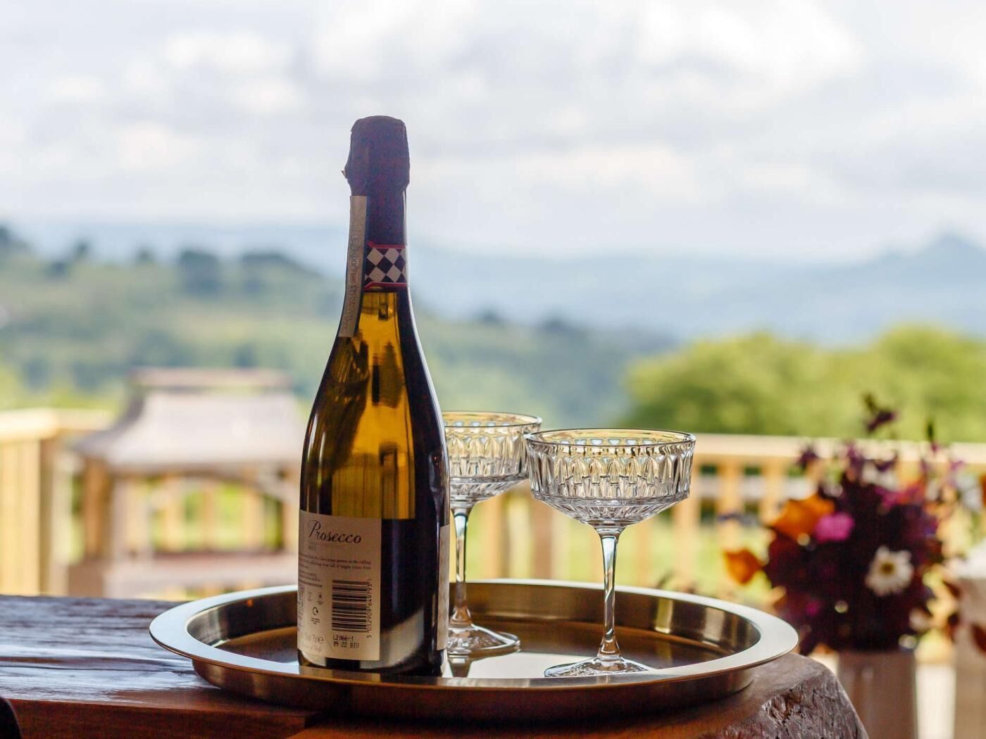 Una bottiglia di prosecco su un vassoio con due calici, con vista su un paesaggio suggestivo.