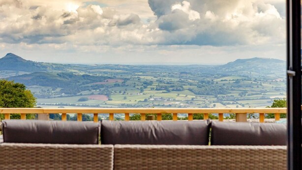 A view from a balcony with a wicker sofa, a wooden railing, and a landscape of fields and hills. - 1 Bed in Abergavenny (oc-88015) (Llanover)