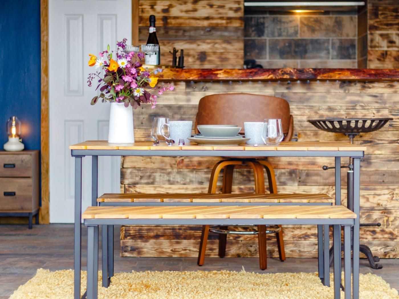 A dining area with a wooden table set for two, a vase of flowers, and a bottle of wine on a rustic wooden counter.