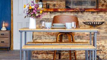 A dining area with a wooden table set for two, a vase of flowers, and a bottle of wine on a rustic wooden counter.