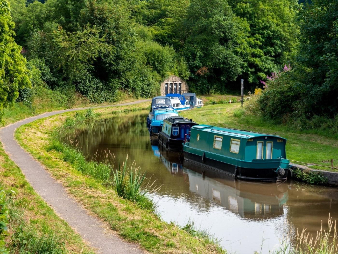 A narrow waterway with boats docked on the right, a pathway on the left, and dense greenery in the background.