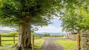 A rural pathway leading to a stone wall and a wooden structure, surrounded by lush greenery and a distant view of hills. - 1 Bed in Abergavenny (oc-88015) (Llanover)
