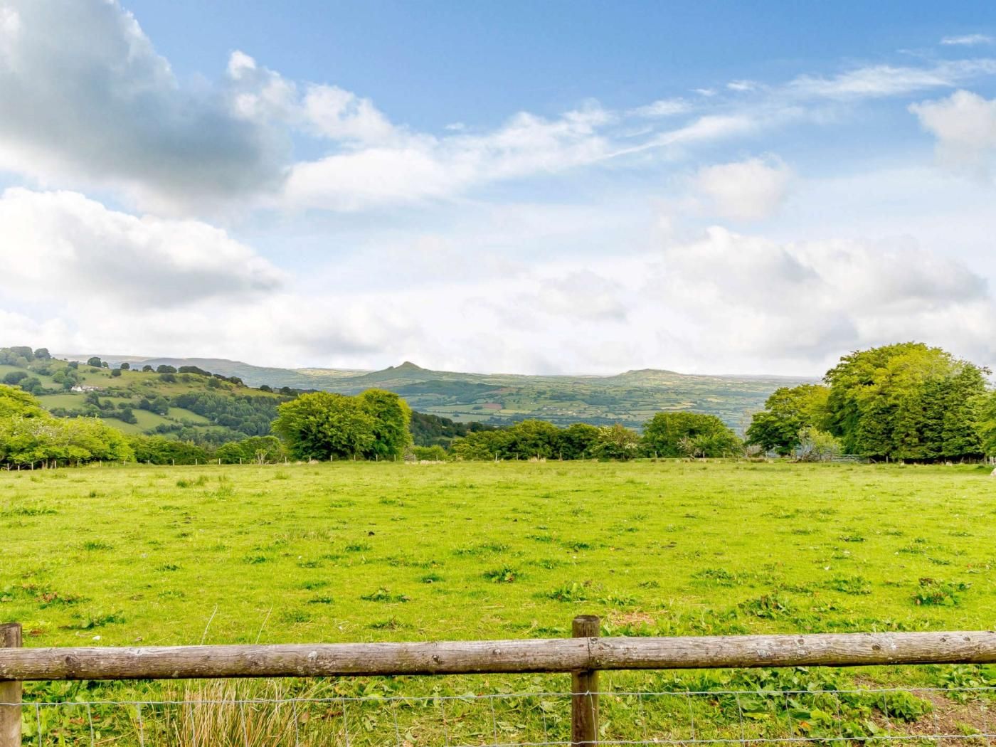A rural landscape with a grassy field, trees, and rolling hills in the background.