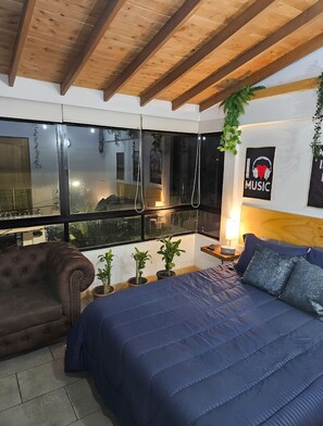 A bedroom with a bed, a brown leather armchair, a bedside table with a lamp, and a wall with a 'I love music' poster. - APARTALOFT - CABAÑA LEON - LAURELES, MEDELLÍN - ANTIOQUIA (Medellín)