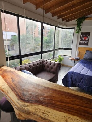 A modern room with a large window, a wooden desk, a tufted sofa, a bed with a blue quilt, and a wall-mounted plant. - APARTALOFT - CABAÑA LEON - LAURELES, MEDELLÍN - ANTIOQUIA (Medellín)