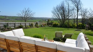 Ein Sitzbereich im Freien mit einer Holzterrasse, einer Couch und zwei Stühlen, mit Blick auf einen gepflegten Rasen und eine weite Landschaft.