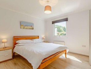 A bedroom with a wooden bed, bedside lamps, a window with a view, and a painting on the wall. - 2 Bed in Saltash (oc-88308) (Notter)